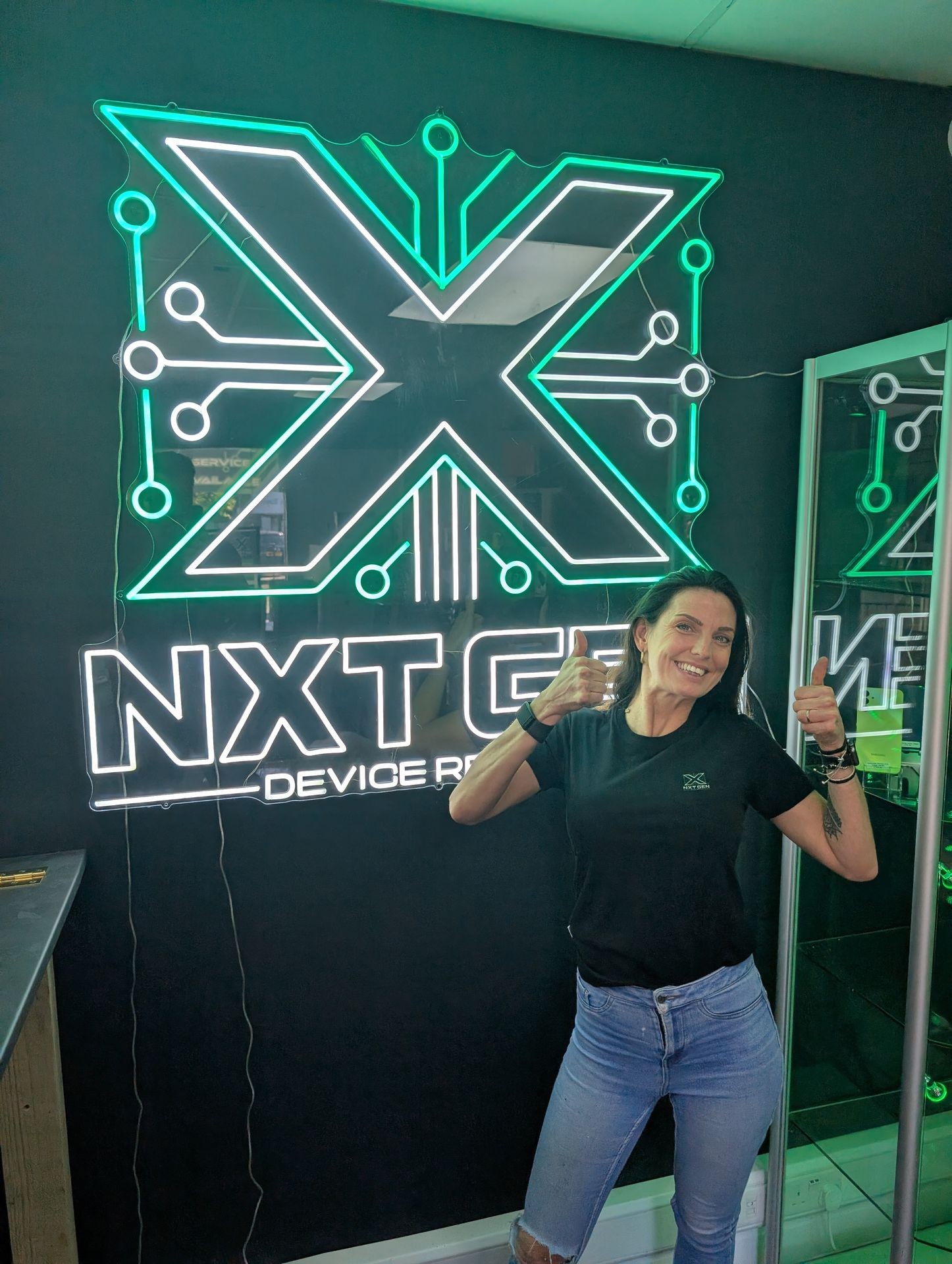 Person giving thumbs up in front of neon logo sign on dark wall.